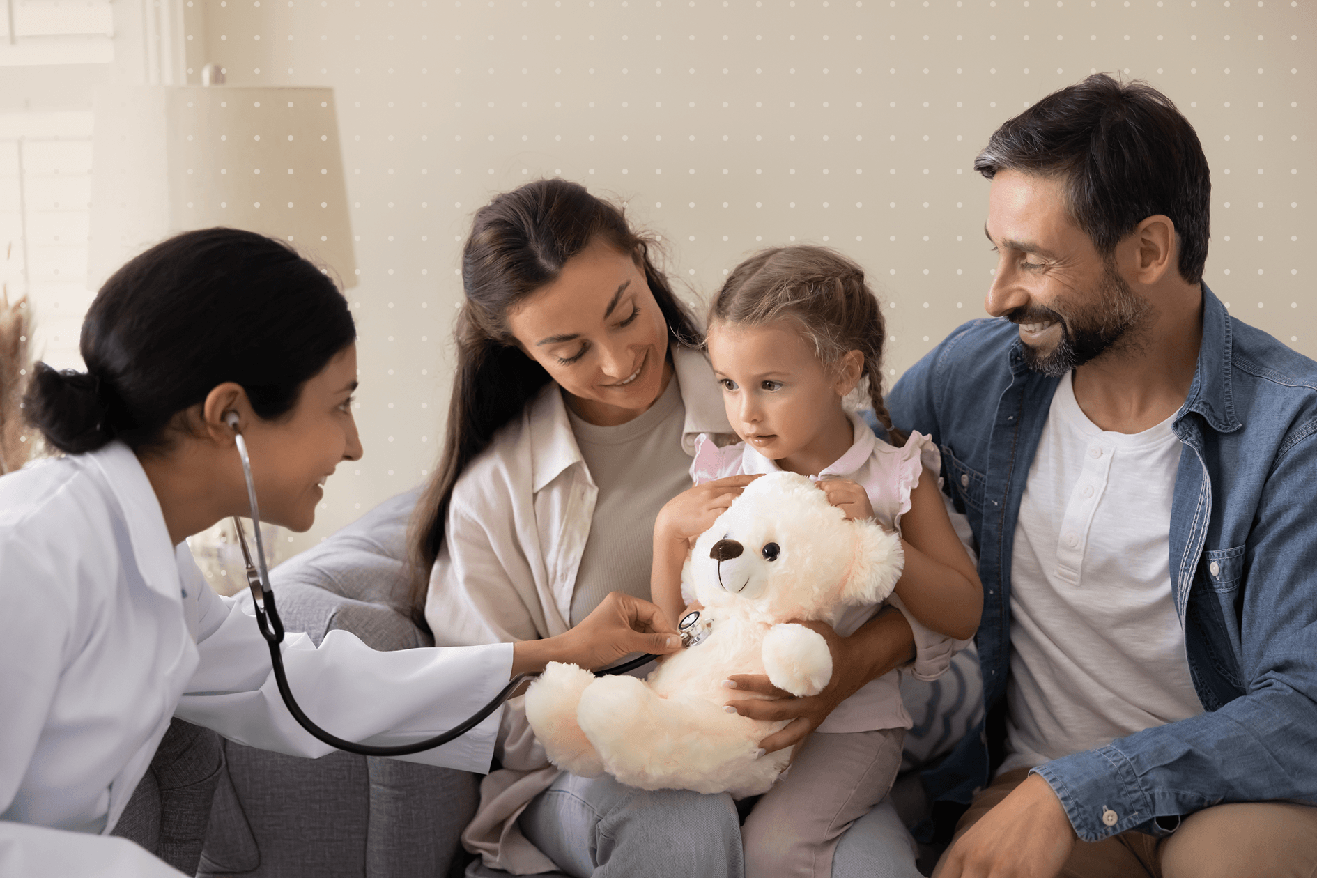 A female doctor on the left with a young family on the right. The doctor is listening to the little girl's heart beat as the girl holds a teddy bear.
